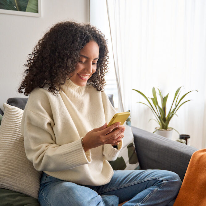 Happy young latin woman sitting on sofa holding mobile phone using cellphone technology doing ecommerce shopping, buying online, texting messages relaxing on couch in cozy living room at home.